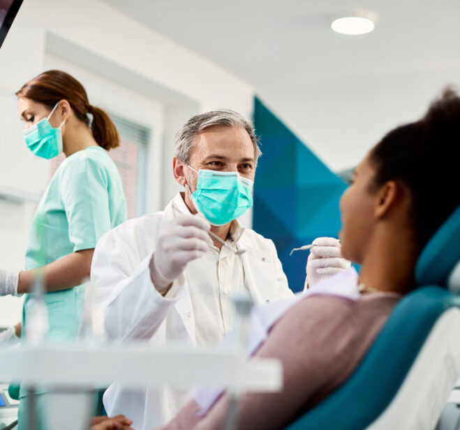 Smiling dentist communicating with African American woman while checking her teeth during dental procedure at dentist's office.